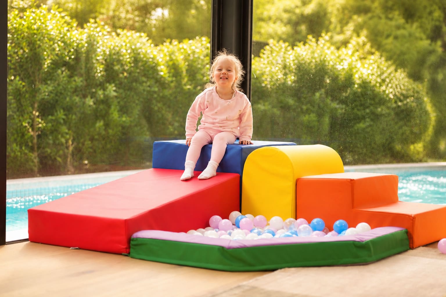 Child sitting on a colorful play structure by a pool with greenery in the background