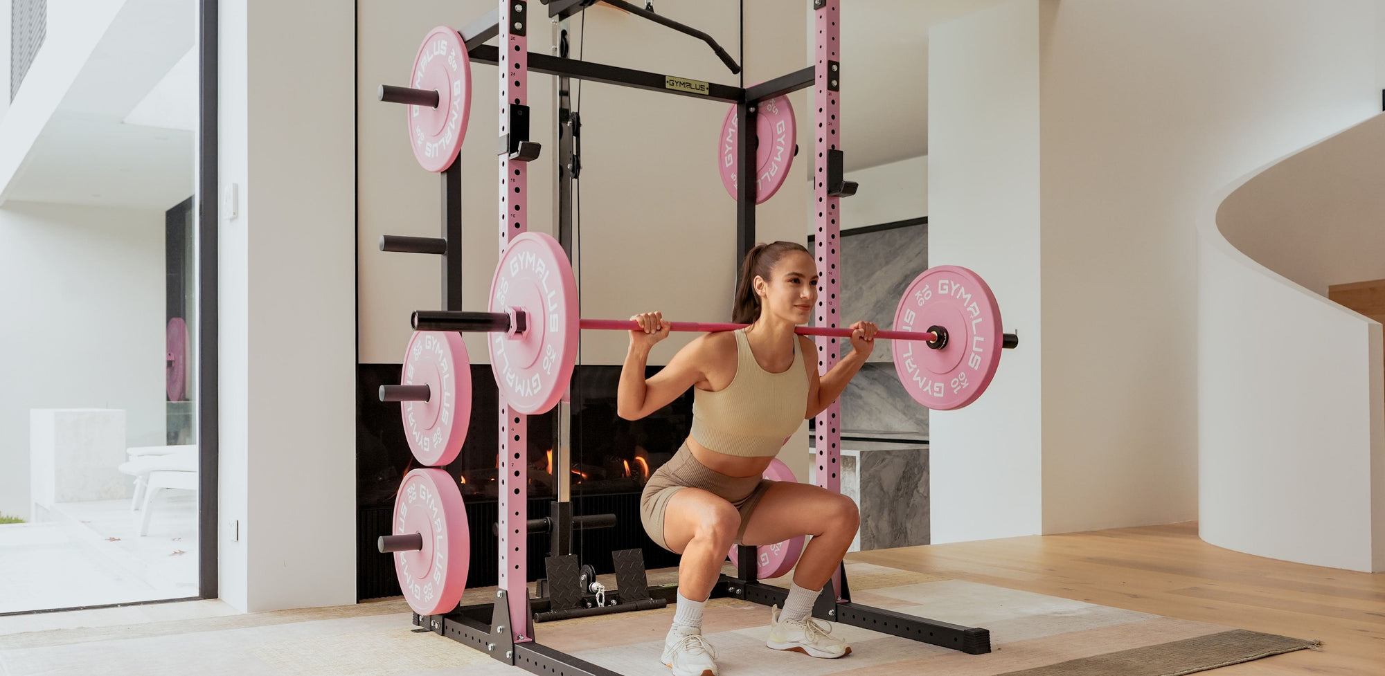 A woman in athletic wear performs a squat inside a pink Gym Plus power rack with pink weight plates in a modern home gym setting.