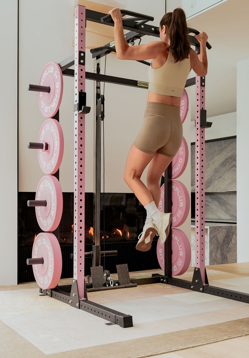 A woman in beige activewear performs a pull-up on a pink and black Gym Plus power rack with pink weight plates in a modern home gym setting.